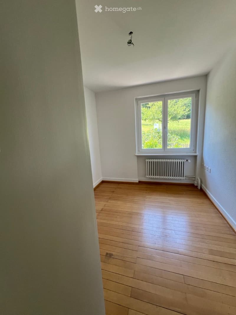 Empty room with wooden flooring, white walls, and a large window with a view of greenery outside. A radiator is visible below the window.