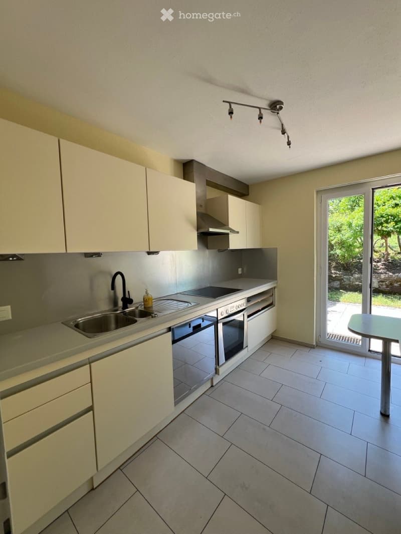 Modern kitchen with light-colored cabinets, stainless steel appliances, and a tiled floor. Features a sink, faucet, oven, and a small table near the sliding glass door leading to an outdoor area.