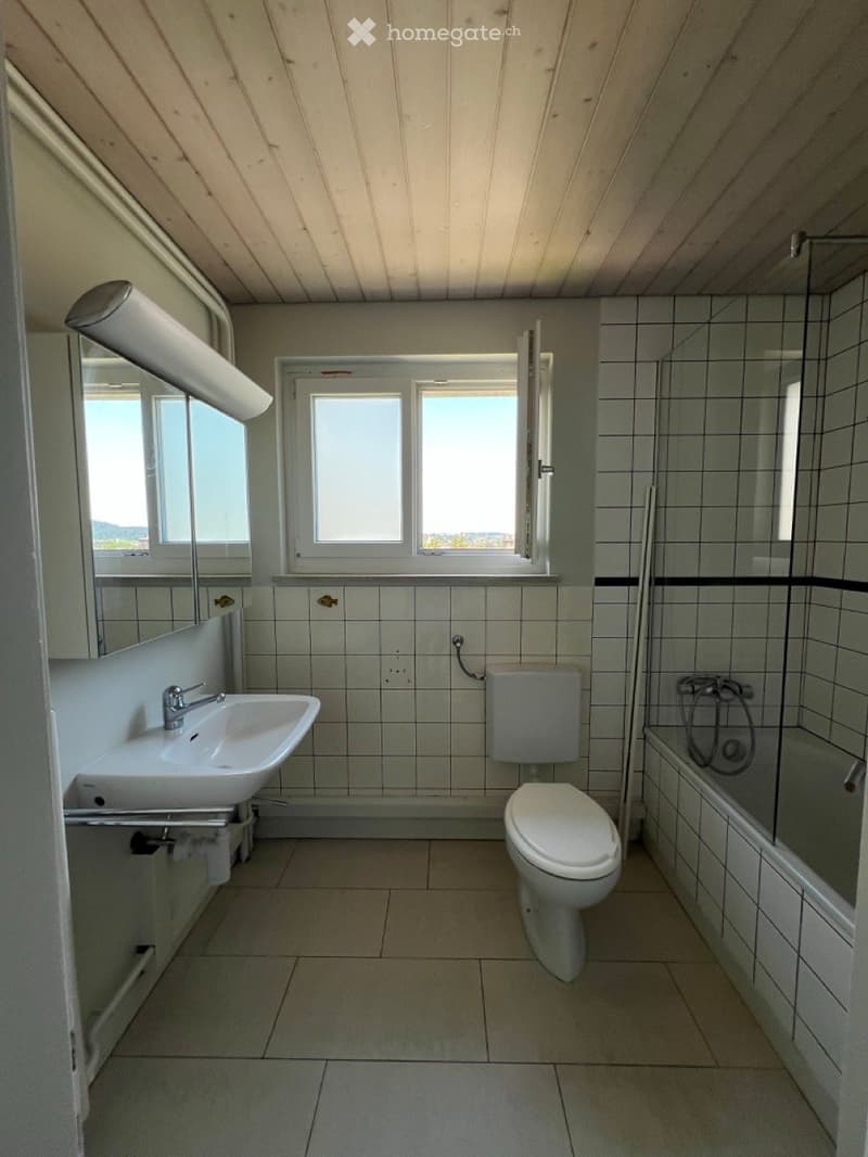 Bathroom with tiled walls and floor, featuring a sink, toilet, bathtub, and a large window providing natural light.