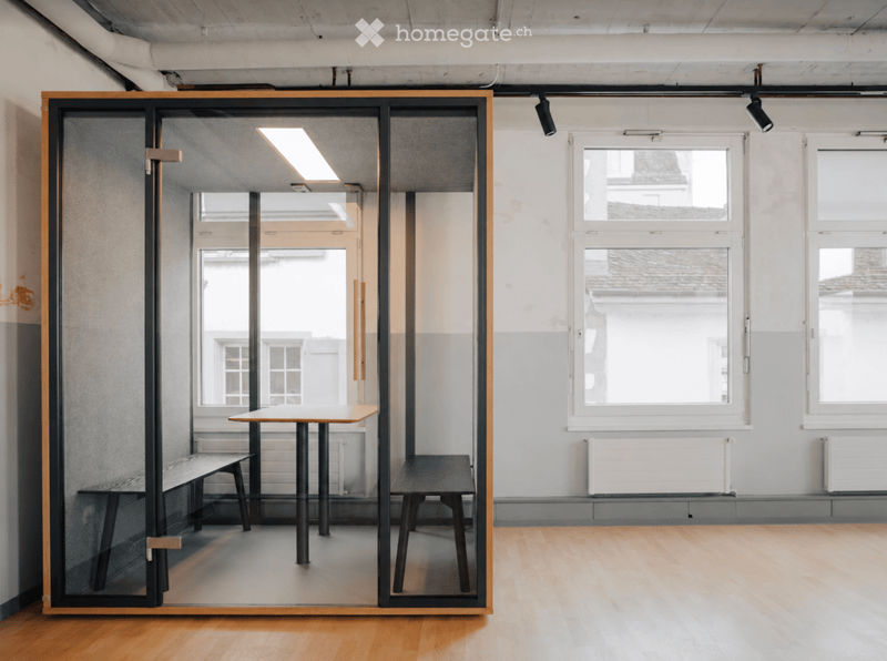 A modern, minimalist room with wooden flooring, large windows, and a glass partition featuring a small table and bench inside.
