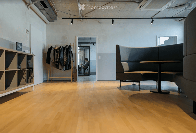 Modern, minimalist room with light wooden flooring, a large L-shaped sofa, a small round table, a wooden shelf, and a coat rack with jackets.