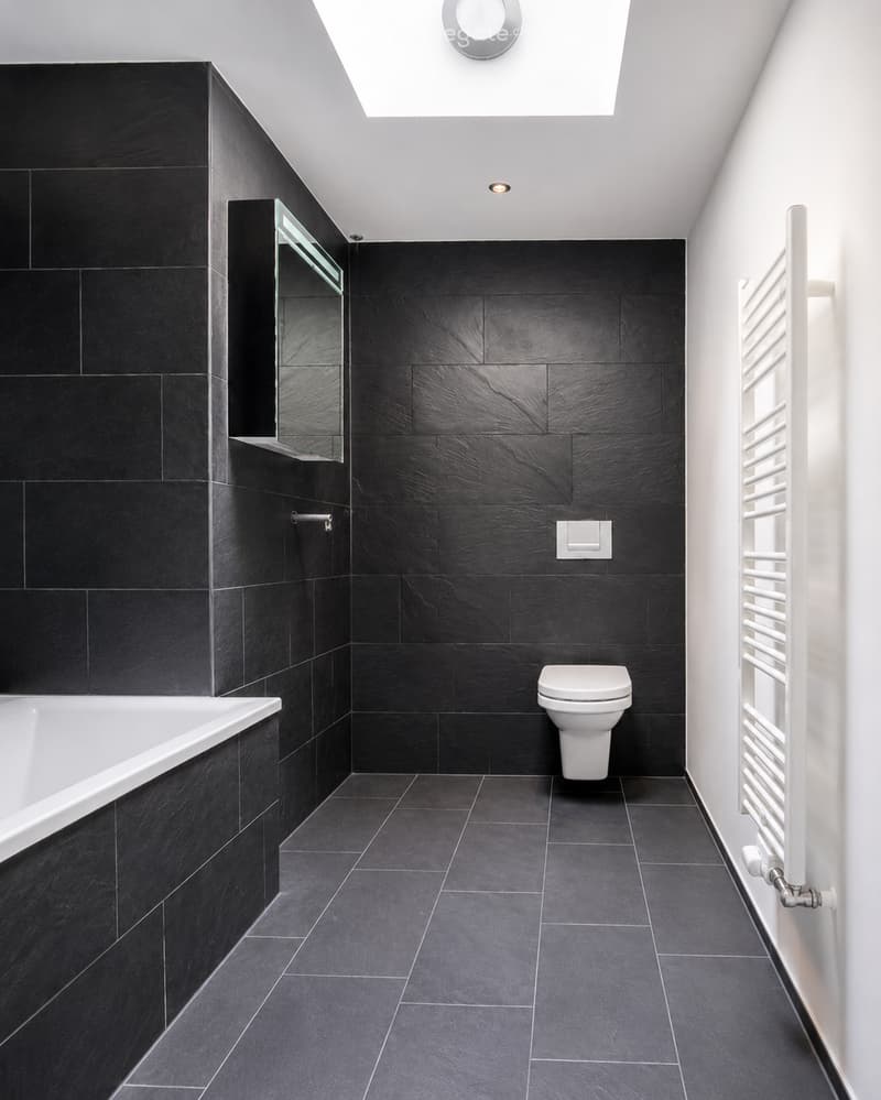 Modern bathroom with black tiles, white bathtub, toilet, mirror, and radiator.