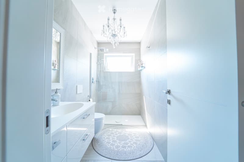 Modern bathroom with white tiles, a large sink with a mirror, a toilet, a shower area with glass enclosure, and a decorative rug on the floor.