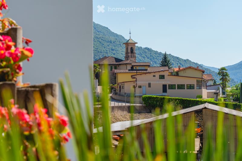 View of a small town with a church tower, surrounded by mountains and greenery, with a modern building and a construction site visible.