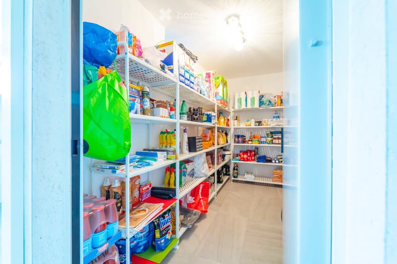 Well-organized pantry with white shelves stocked with cleaning supplies, food items, and plastic bags. The room features light wooden flooring and white walls.