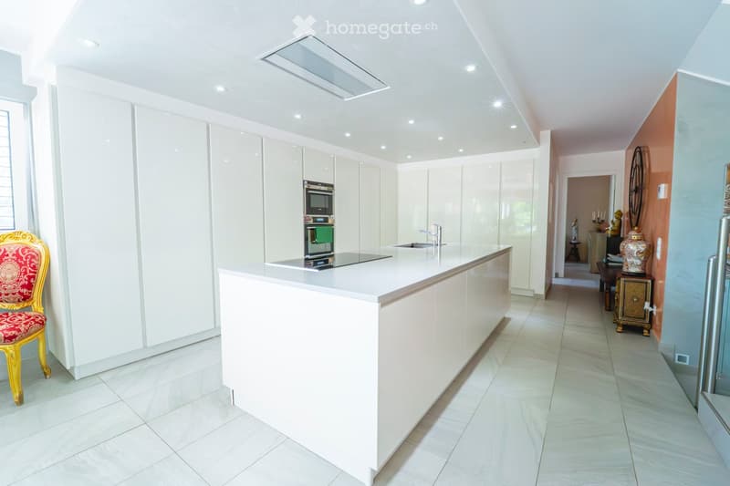 Modern hallway leading to a kitchen area with white cabinets, a central island, and a decorative chair near the entrance.