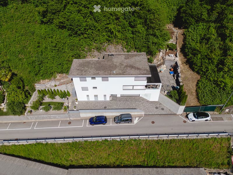 Aerial view of a two-story white house with a gray roof, surrounded by greenery and a parking area with three cars.