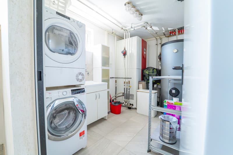 A modern bathroom with a washing machine, dishwasher, water heater, and cabinet. The space is well-lit with white walls and tiled flooring.