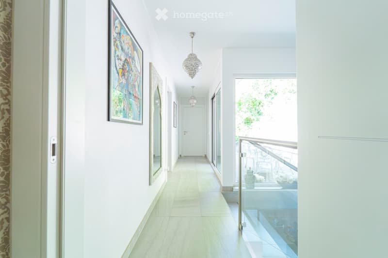 A modern, well-lit hallway with white walls, tiled flooring, and a chandelier hanging from the ceiling. The hallway features framed artwork on the walls and leads to a balcony area with a glass railing.
