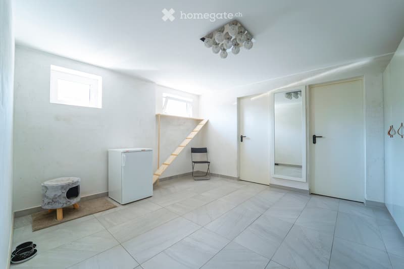 A modern, minimalist room with white tiled flooring, white walls, and a skylight. Features include a small refrigerator, a wooden staircase leading to an upper level, a black folding chair, a fur-covered stool, and a large mirror on the wall.