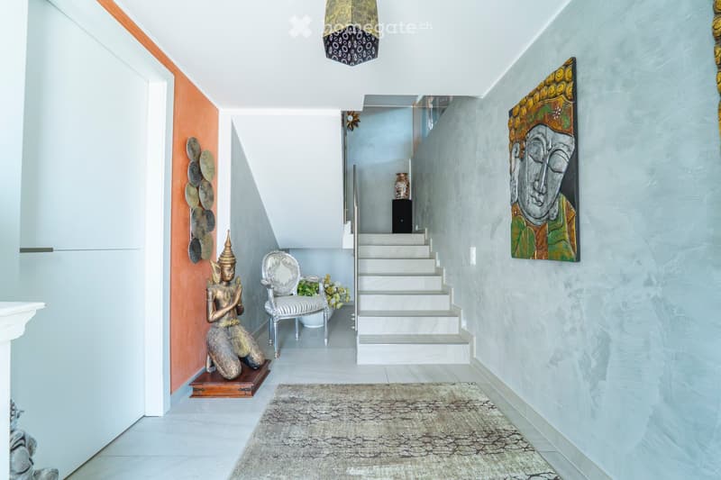 Modern hallway with light blue walls, white staircase, decorative artwork, a small chair, and a statue on a wooden base.