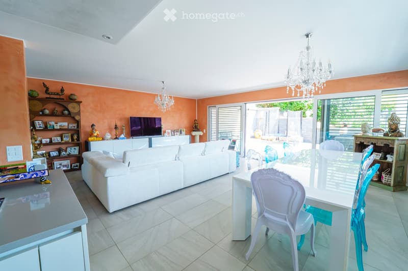 Bright living room with white sofa, dining table, chairs, TV, shelves, and chandeliers. Large glass doors open to a patio.