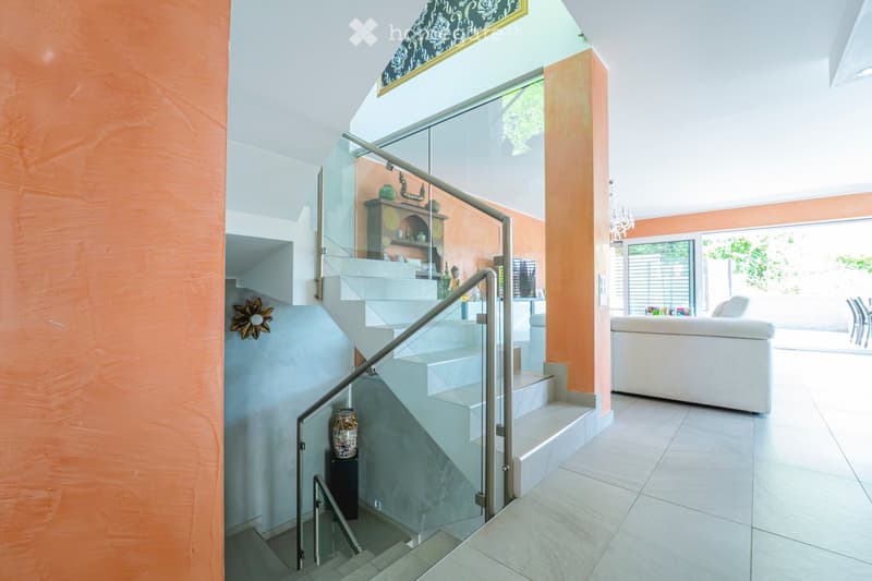 Modern interior with a staircase featuring glass railings, a white sofa, a dining table with chairs, and decorative shelves with vases.