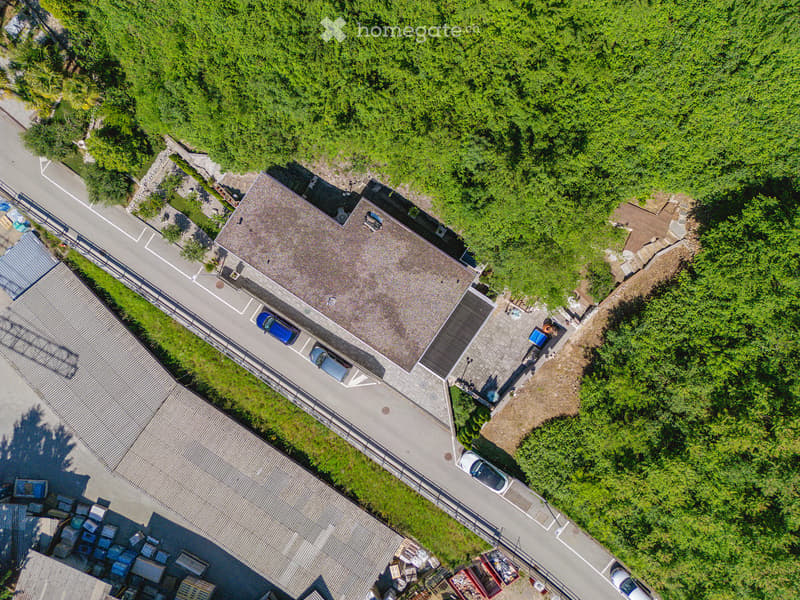 Aerial view of a two-story building with a gray roof, surrounded by greenery and a parking area with several cars.