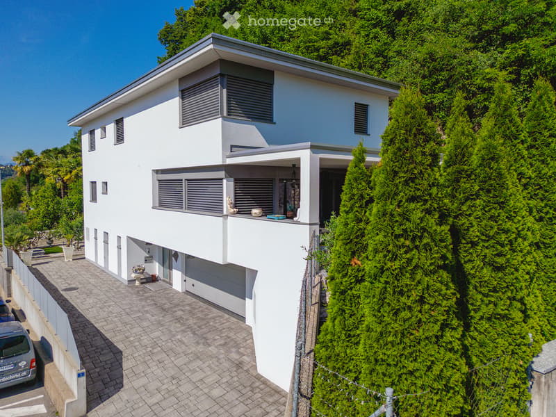 Modern two-story white house with large windows, grey shutters, and a balcony. The house is surrounded by lush greenery and has a paved driveway leading to a garage.