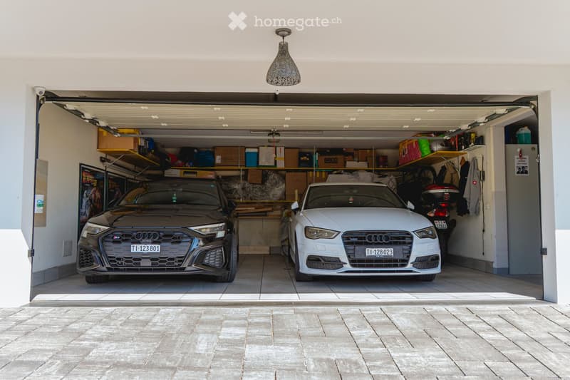 Garage with two Audi cars parked inside, shelves with boxes and items, and a hanging light fixture.