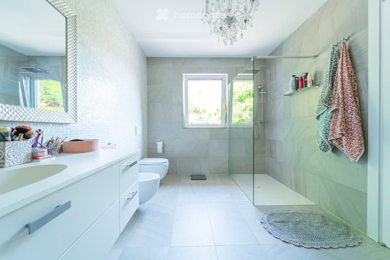 Modern bathroom with light grey tiles, a large sink, a bathtub, a toilet, a shower area, a chandelier, and a decorative rug.