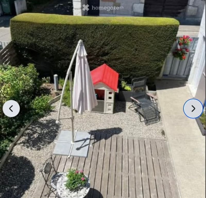 A small, well-maintained garden area with a wooden deck, patio furniture, a playhouse, and a large umbrella. The space is bordered by a neatly trimmed hedge and features potted plants.