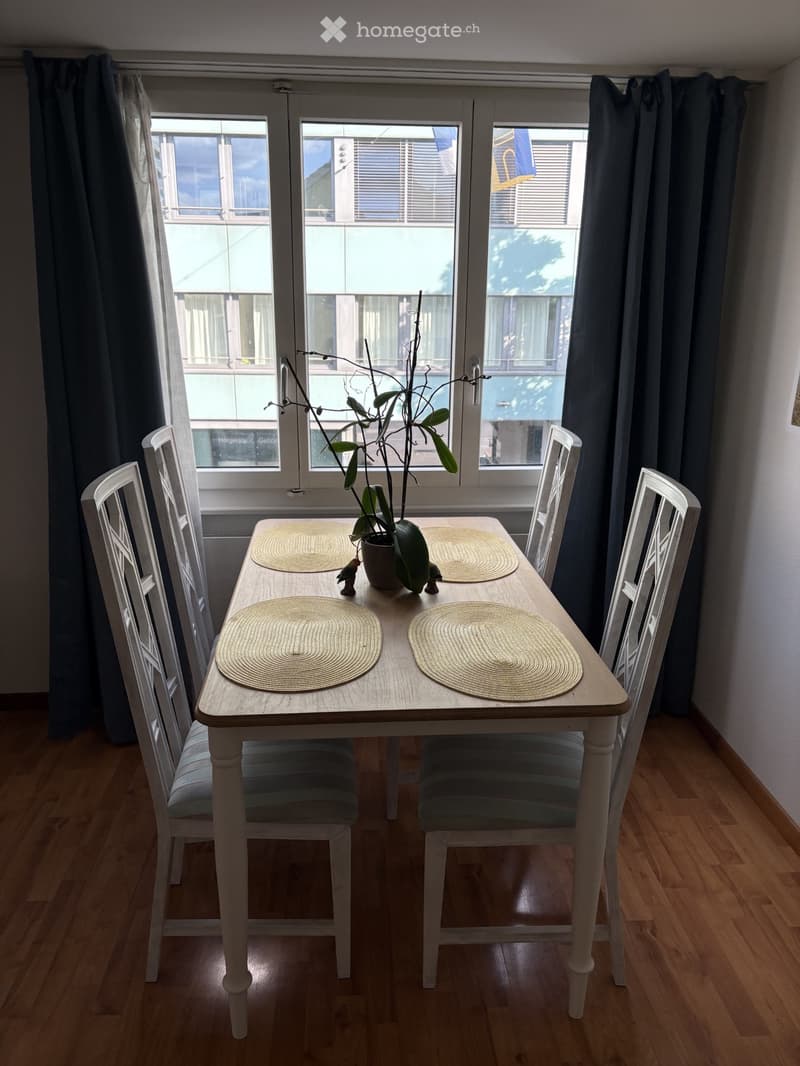 Dining area with a wooden table, four chairs, placemats, and a potted plant near a large window with blinds.