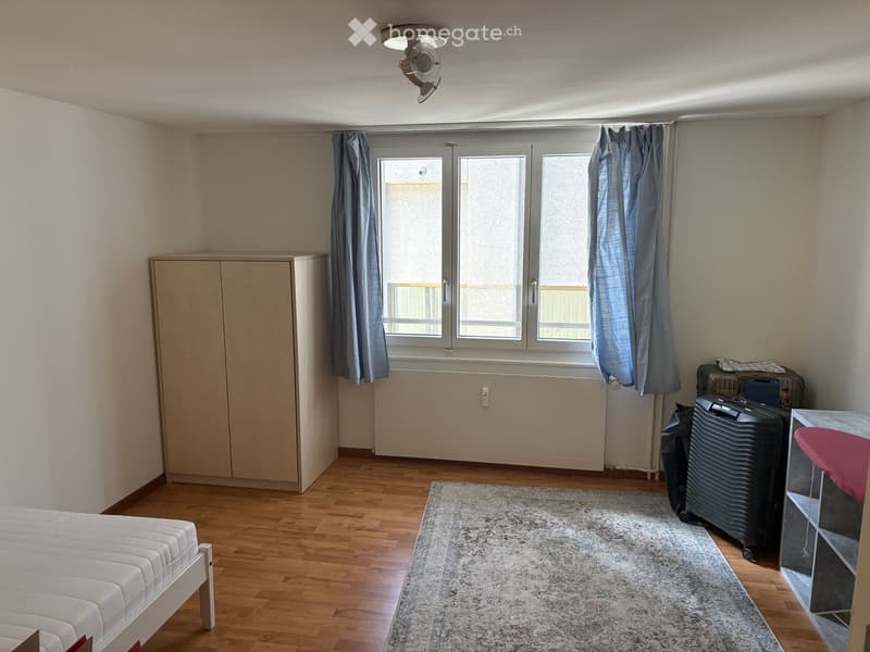 A furnished bedroom with a bed, wardrobe, suitcase, table, chair, and curtains. The room features wooden flooring and a large window with a view.