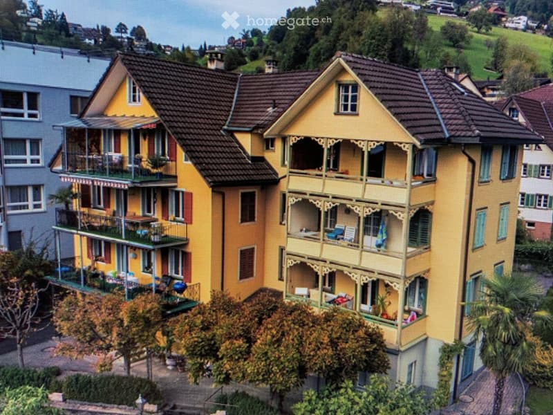 A multi-story yellow building with balconies and ornate architectural details, surrounded by greenery and neighboring structures.