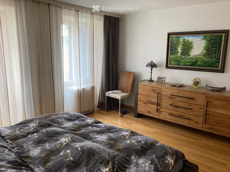 Cozy bedroom with wooden flooring, a large bed with patterned bedding, a wooden dresser with drawers, a chair, and a decorative lamp on the dresser.