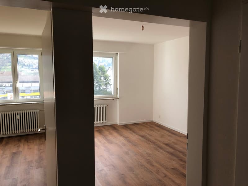 Empty room with wooden flooring, white walls, and large windows offering a view of the outside area.