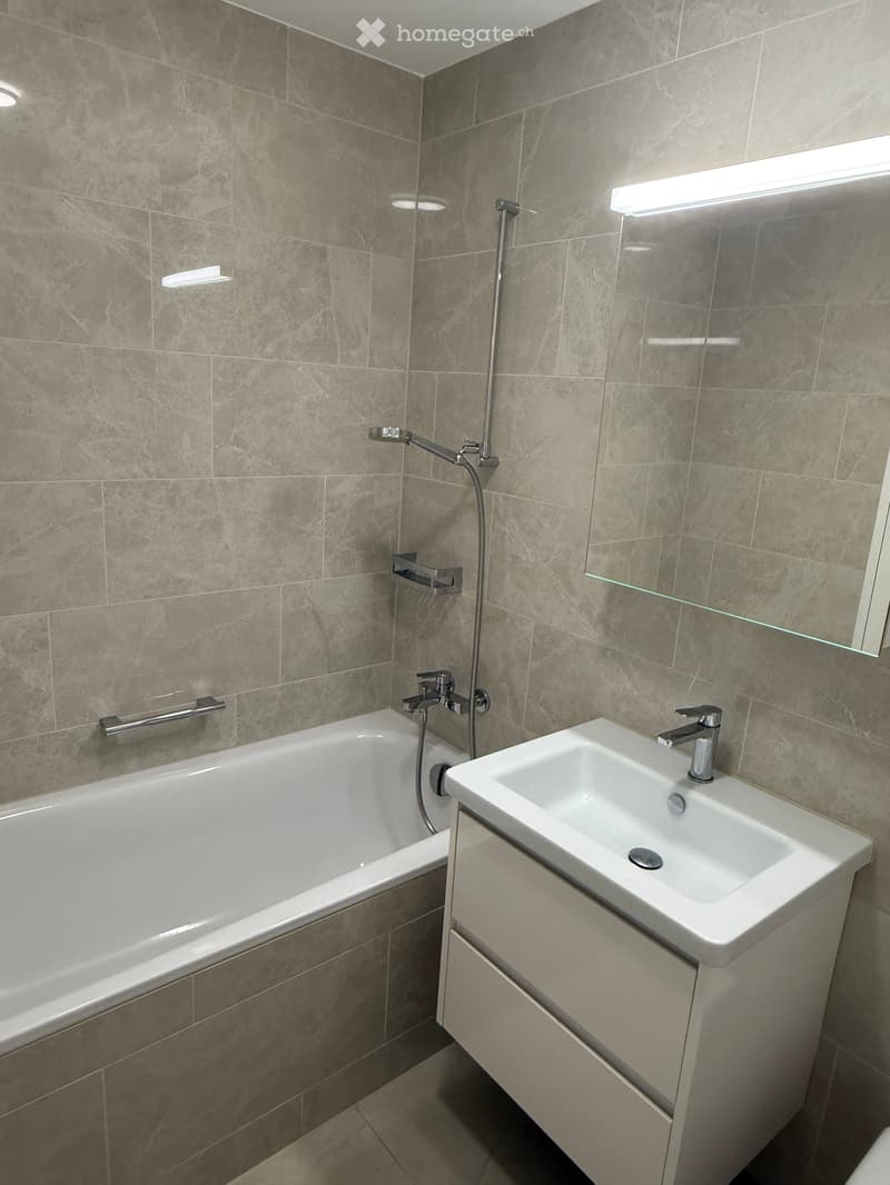 Modern bathroom with a bathtub, sink, and showerhead. Features grey tiled walls and a built-in vanity cabinet.