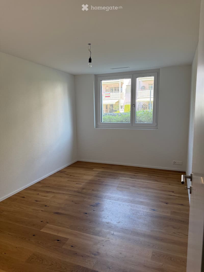 Empty room with wooden flooring, white walls, and a large window with a view of the outside. The room has a single light fixture hanging from the ceiling and a door visible on the right side.