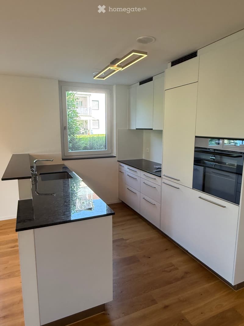 Modern kitchen with white cabinets, black countertops, a sink with a faucet, and an oven. The space features wooden flooring and a window with a view of greenery.