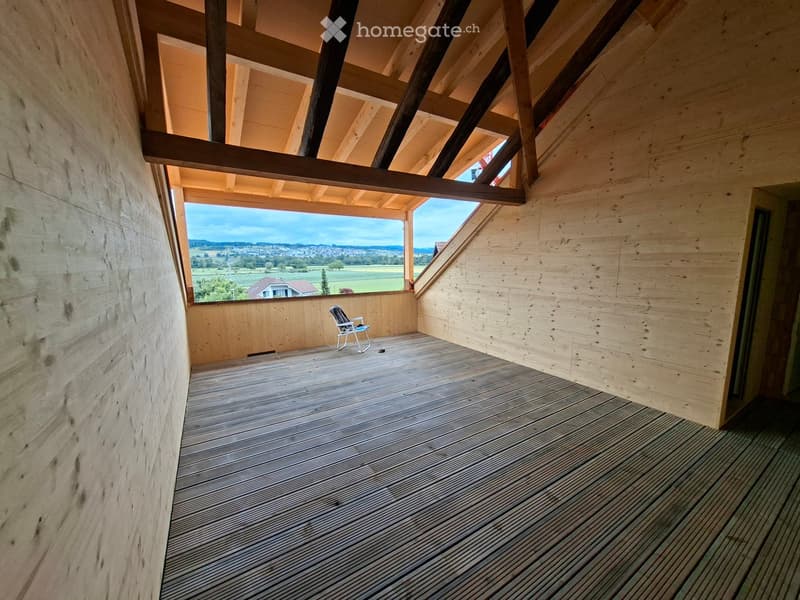 Wooden balcony with slatted floor, exposed wooden beams, and a scenic countryside view through large windows.
