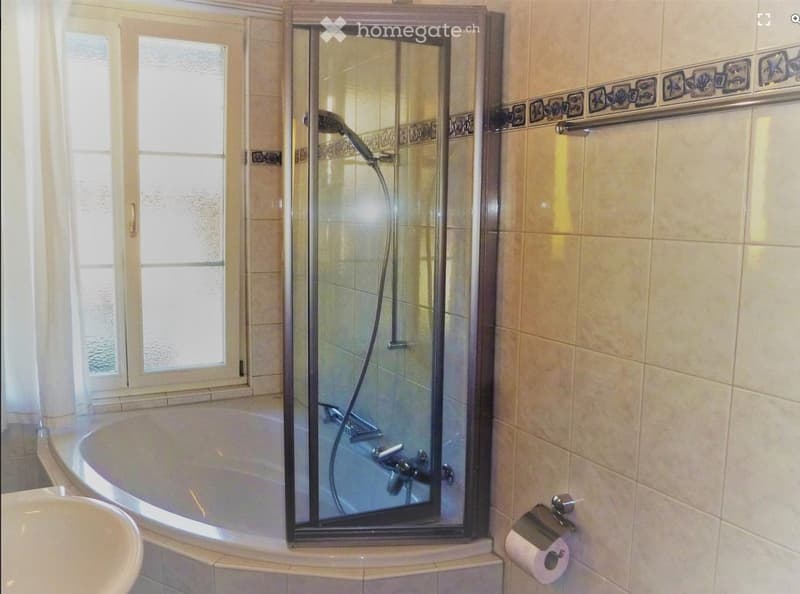 Bathroom with a bathtub, glass-enclosed shower, sink, and toilet paper holder. The walls are tiled with a decorative border near the ceiling.