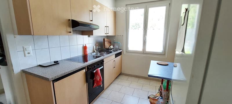 A modern kitchen with light wooden cabinets, a tiled backsplash, and a granite countertop. Features include an electric stove, sink, and a small table with a cutting board.