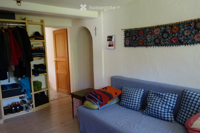A cozy living room with a grey sofa, multiple pillows, a small wooden table, and a wooden wardrobe filled with clothes and shoes.