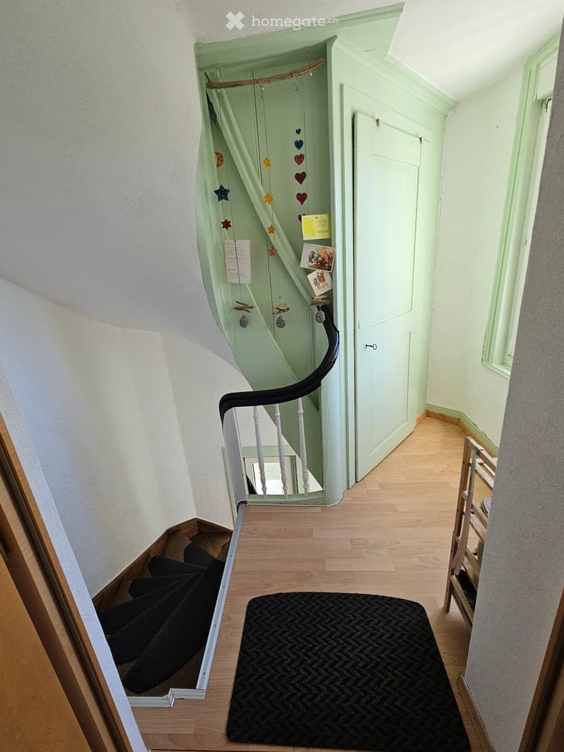 A narrow hallway with light green walls, wooden flooring, and a staircase leading upwards. A black runner rug is placed on the floor, and a small wooden chair is visible to the right. The staircase features a white railing and a black handrail, with a dec