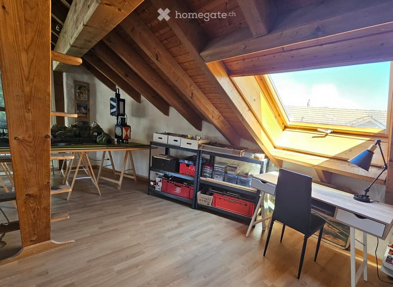 Bright attic workspace with wooden floors, slanted wooden ceiling, large skylight window, and various furniture including a desk, chair, shelves, and storage boxes.