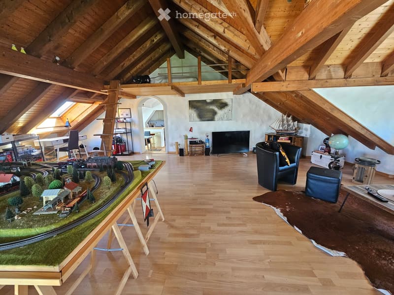 Spacious attic living room with wooden beams, hardwood floors, a model train set, a sofa, a television, and a model ship on a shelf.