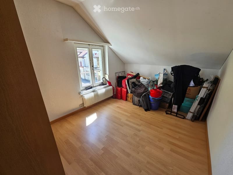 An attic room with wooden flooring, slanted ceilings, and a window offering a view of the roof and surrounding area. The room is empty, with no furniture or furnishings.