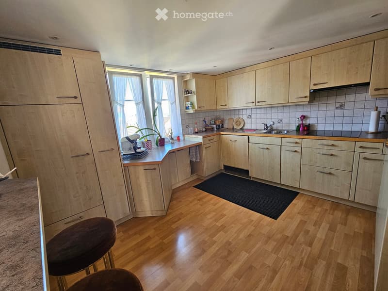 The kitchen features light wooden cabinets, a central island with a countertop, and a sink area with a faucet. There are two stools near the island, and the floor is made of light wooden planks. The kitchen is well-lit with natural light coming through th
