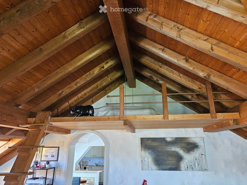 Loft-style room with exposed wooden beams and a slanted ceiling. Features a shelf with a decorative statue, a mirror, a framed picture, a small table with a chair, and a visible kitchen area with a counter, sink, and faucet.