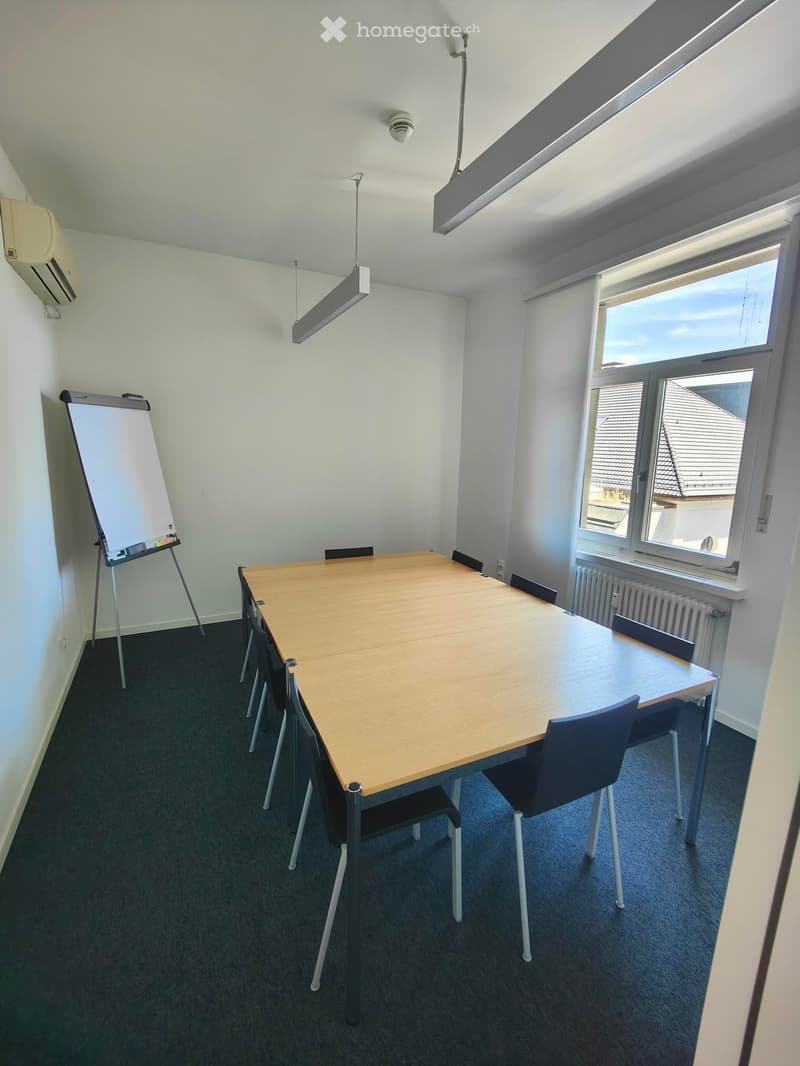 A modern meeting room with a large wooden table, several chairs, a flipchart on a stand, and large windows with a view of the roof.