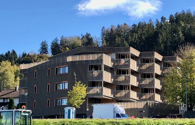 Modern apartment building with balconies, brown exterior, multiple floors, surrounded by trees and grass