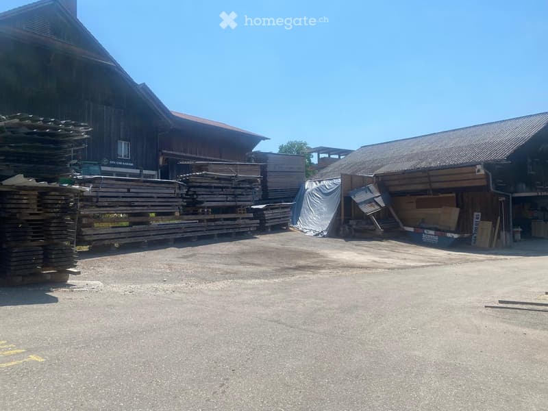 Outdoor view of a large wooden building with a garage and stacks of wooden planks and materials nearby.