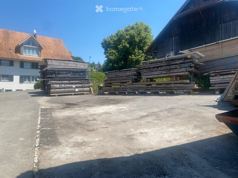 Outdoor area with a concrete floor, stacks of wooden planks, and a large wooden building in the background.