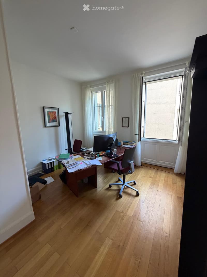 Office space with wooden flooring, large window with curtains, desk with office supplies, two chairs, and a framed picture on the wall.