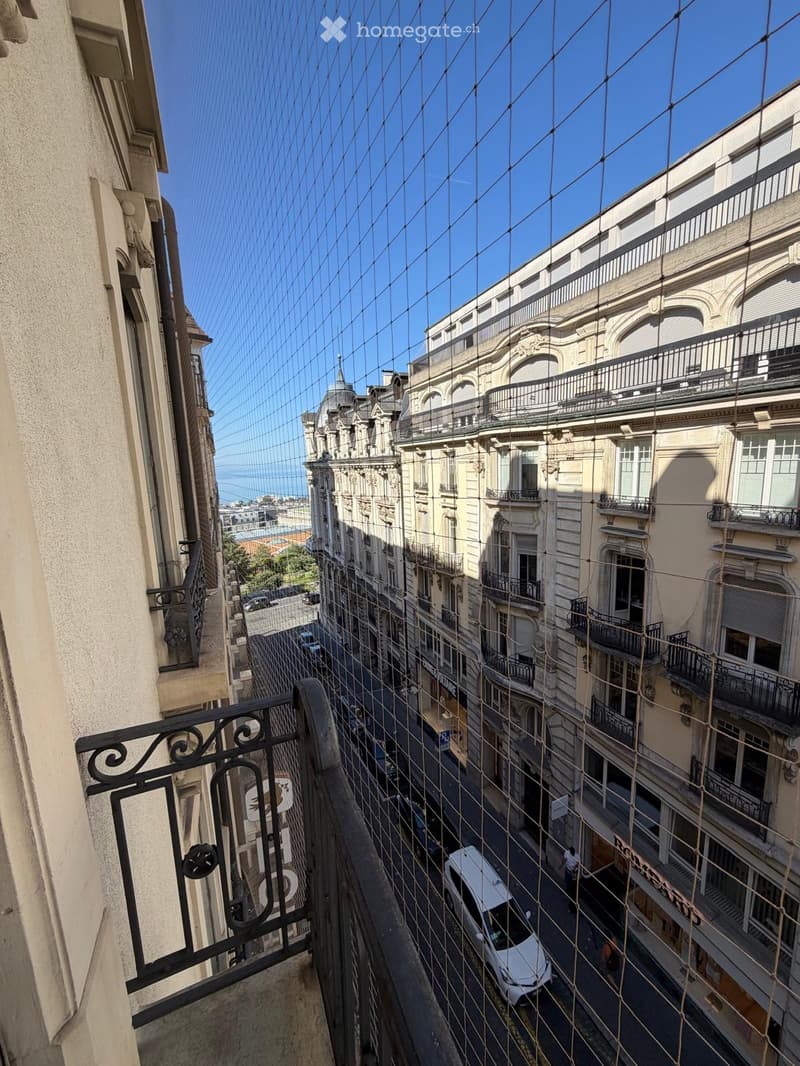 Balcony overlooking a narrow street lined with multi-story buildings, featuring ornate balconies and railings. A white van is parked on the street below.