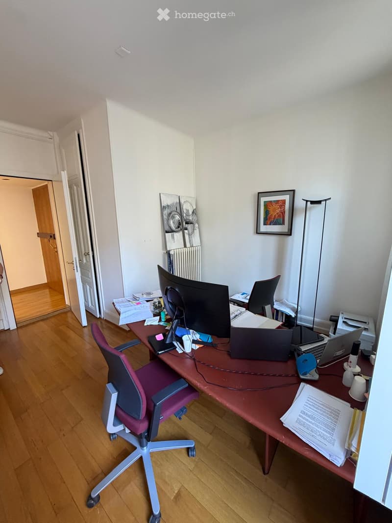 A furnished room with a wooden floor, a large desk with a laptop, monitor, and office supplies, a purple office chair, and a framed picture on the wall.