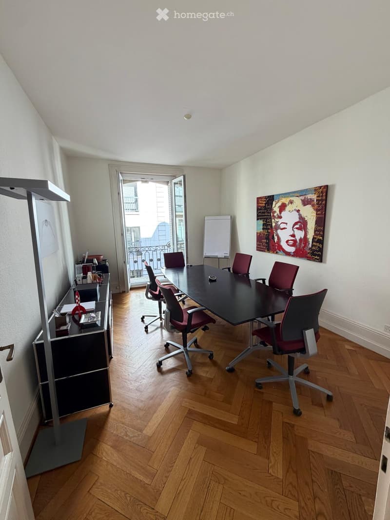Office space with wooden flooring, a large black conference table surrounded by red chairs, a cabinet with office supplies, and a large screen on the wall. An open door leads to a balcony.