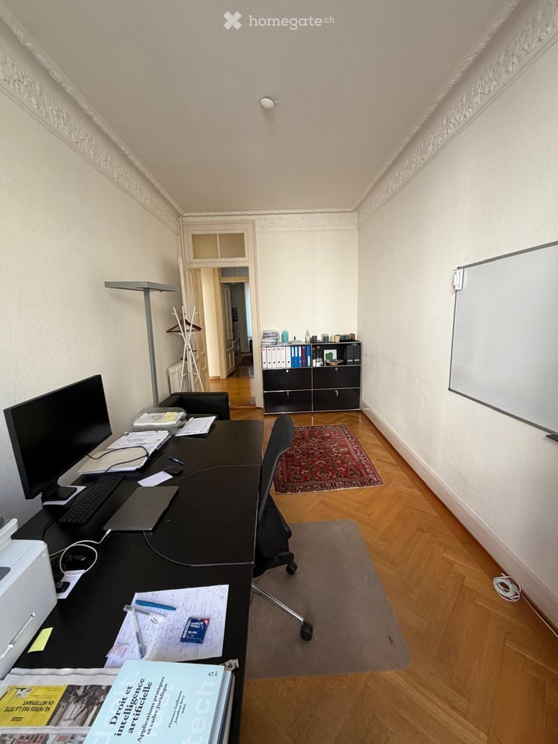 Office space with a wooden floor, a large desk with a laptop, keyboard, and various office supplies, a chair, a whiteboard, a cabinet with books and files, and a small rug.