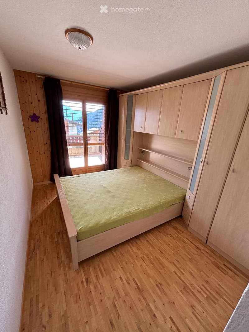 Bedroom with wooden flooring, a single bed with green bedding, built-in wardrobe and cabinets, large window with balcony access, and wooden door.